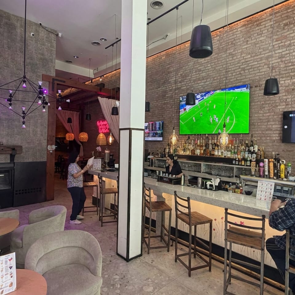 Modern restaurant bar with exposed brick walls, pendant lights, televisions showing sports, and patrons seated at tables and counter.