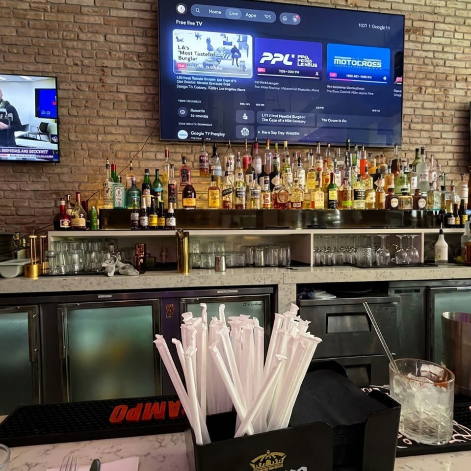 Restaurant bar lined with liquor bottles, televisions, cocktail menu, glassware, mixers, and a container of white straws.
